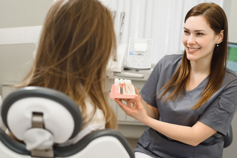 a happy dental assistant showing a patient a dental implant model so she can explain how dental implants will improve her smile.