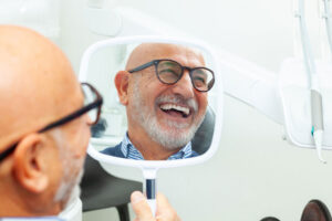 Smiling senior man with glasses admiring his new dental work in a handheld mirror at a dental clinic, reflecting satisfaction and confidence after treatment.