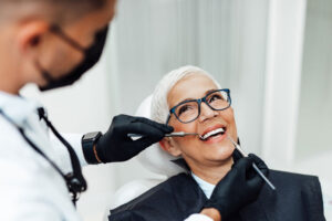 Smiling senior woman with short white hair and glasses receiving a dental checkup from a masked dentist wearing black gloves, showcasing a confident and healthy smile during her appointment.