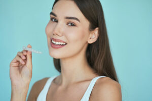 Smiling woman holding a clear orthodontic aligner near her mouth against a light blue background.