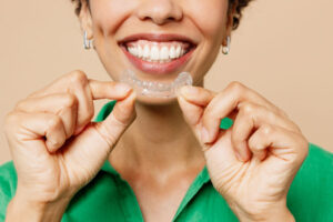 Close-up of a smiling woman in a green shirt holding a clear orthodontic aligner in front of her mouth.