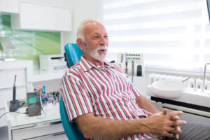 Older man with white hair and beard sits relaxed on a teal dental chair in a bright, modern dental office, wearing a red and white striped shirt, with dental equipment and sunlight in the background.
