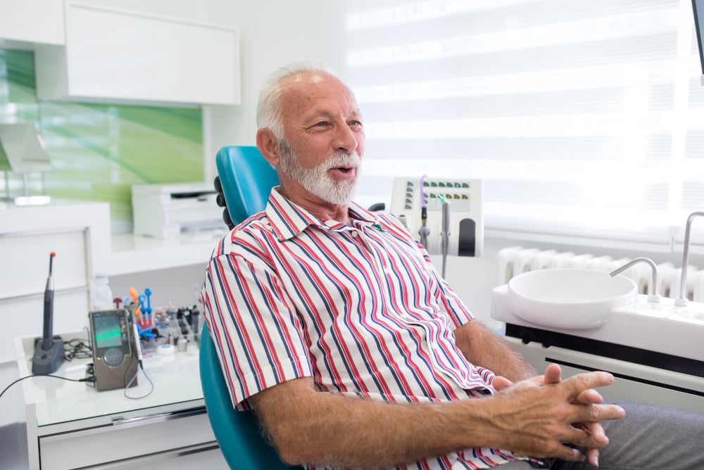 Older man with white hair and beard sits relaxed on a teal dental chair in a bright, modern dental office, wearing a red and white striped shirt, with dental equipment and sunlight in the background.