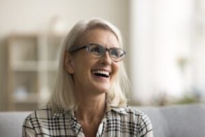 Smiling older woman with shoulder-length blonde hair and glasses, wearing a black-and-white plaid shirt. She is sitting indoors on a light-colored couch, with a blurred shelf and soft light behind her.