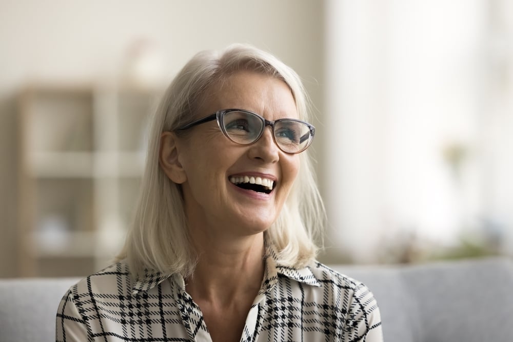 Smiling older woman with shoulder-length blonde hair and glasses, wearing a black-and-white plaid shirt. She is sitting indoors on a light-colored couch, with a blurred shelf and soft light behind her.