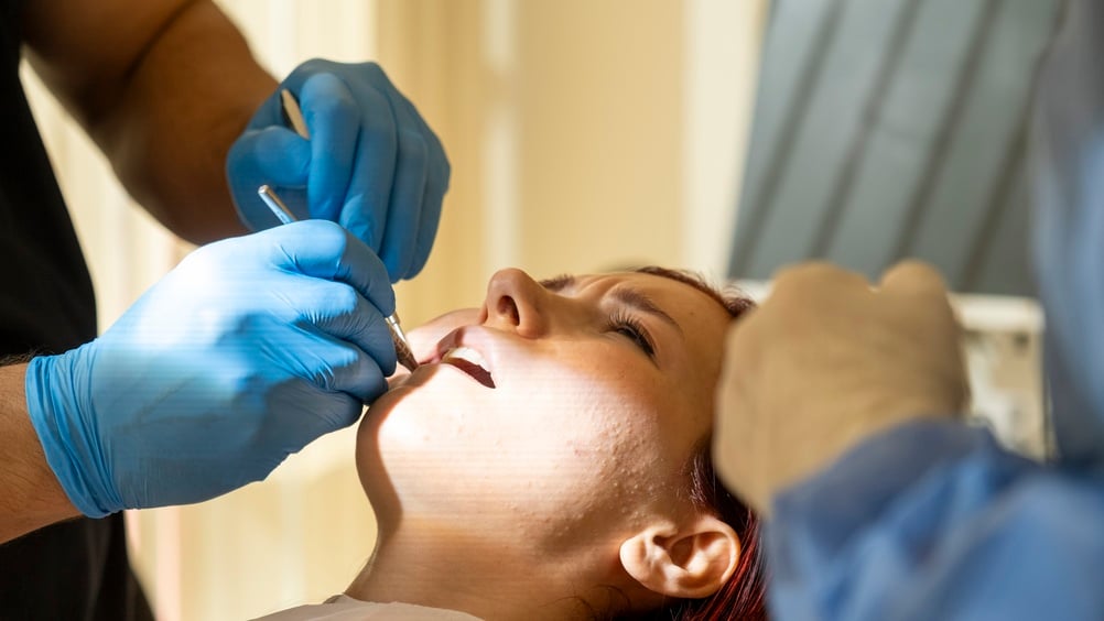 A person reclines in a dental chair with mouth open as a dentist wearing blue gloves works on their teeth using dental tools. Light illuminates the patient’s face, drawing attention to the ongoing dental procedure.