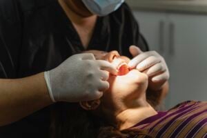 A dentist in gloves and a mask examines a patient’s mouth. The patient reclines in the dental chair, wearing a striped shirt. Bright light highlights the scene, focusing on the patient’s open mouth.